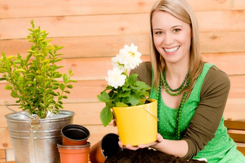 A woman with long blonde hair smiling at the camera while holding a vibrant yellow flower pot containing a flowering plant with large white blossoms and green foliage, situated on a wooden outdoor surface. Behind her is a light wooden fence, and to her left are gardening items including a tree in a metallic pot, a small empty terracotta pot, and a black plastic pot. The background suggests a well-maintained garden or patio area with natural lighting indicating a bright day, and the scene reflects general gardening activities and outdoor plant care, consistent with professional garden maintenance services in Hayes, UK.