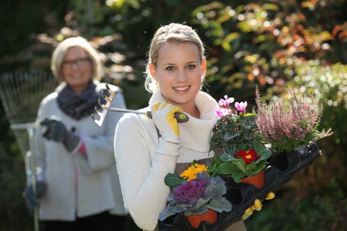 A young woman with blonde hair and a friendly expression is standing outdoors in a garden during daylight, holding a tray containing a variety of potted flowers with vibrant purple, pink, red, and yellow blooms. She is dressed in a white sweater and yellow gardening gloves, appearing prepared for outdoor gardening tasks. Behind her, an older woman with glasses and a scarf is smiling and holding a garden rake, also dressed for gardening in a light jacket. The garden features a well-maintained lawn in the foreground with lush, green grass, and a backdrop of colorful foliage, shrubs, and trees indicating a landscaped outdoor space in Hayes, with natural lighting suggesting a clear, sunny day. This scene illustrates a gardening activity, aligned with outdoor lawn and garden care services offered by Hedge Trimming Hayes, emphasizing local landscaping and garden maintenance in the area.
